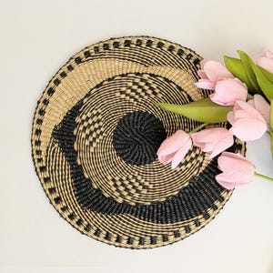 Puede incluir: Una cesta redonda tejida con un diseño en espiral en beige natural y negro. La cesta está decorada con un patrón de puntos alrededor del borde. Tulipanes rosas con tallos y hojas verdes están a la derecha de la cesta.