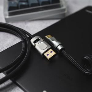 May include: A close-up of a black braided cable with a silver USB-C connector on one end and a silver USB-A connector on the other end. The cable is coiled and resting on a black surface with the word "iPad" printed on it.