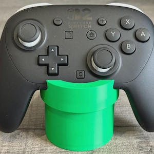 May include: A black Nintendo Switch Pro Controller with white accents, resting on a bright green stand. The controller has buttons labeled A, B, X, and Y, and a directional pad. The stand resembles a green pipe.