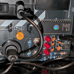 May include: A black circuit board featuring a central USB port with a connected black cable, three red LED lights, various orange disc capacitors, blue resistors, two silver inductors labeled 102, and an STMicroelectronics M27C256B chip.