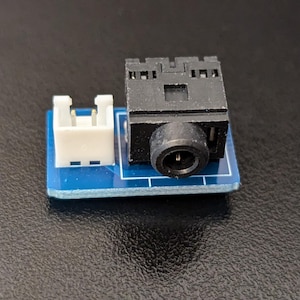 Könnte beinhalten: Eine kleine blaue Platine mit einer schwarzen Audiobuchse und einem weißen Stecker.