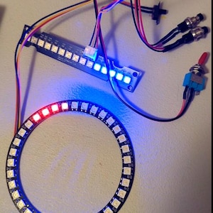 Op de afbeelding: Een verlichte LED-printplaat met een cirkelvormige ring van lampjes, draden en een schakelaar. De ring is verlicht met rode en blauwe lampjes. Een rechthoekige printplaat met meerdere LED-lampjes is ook zichtbaar.