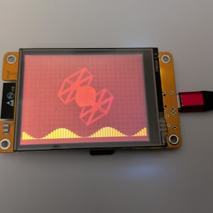 Puede incluir: Un pequeño dispositivo electrónico con una pantalla que muestra un gráfico rojo de un caza TIE y una onda de sonido amarilla. El dispositivo tiene una placa de circuito amarilla y está conectado a un cable negro con un conector rojo.