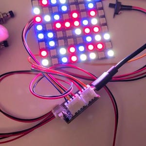 May include: An electronic LED matrix display board with a grid of illuminated red, blue, and white lights. The square circuit board is connected via multi-colored wires to a small controller module and a metal toggle switch component.