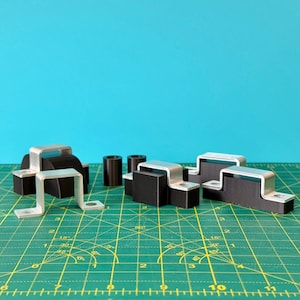 Puede incluir: Un surtido de componentes de goma negra y metal plateado, incluyendo soportes y piezas cilíndricas, se disponen sobre una superficie de rejilla verde contra un fondo turquesa. Los componentes parecen ser piezas industriales o mecánicas.