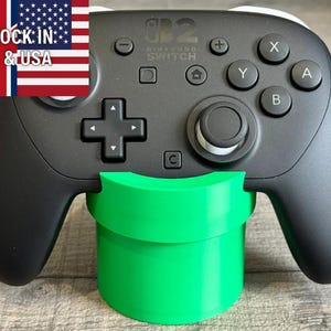 May include: Black Nintendo Switch Pro Controller with green pipe-shaped stand. The controller has buttons labeled X, Y, A, and B, and a directional pad. The stand is a bright green color. The image also includes the text "STOCK IN UK & USA".