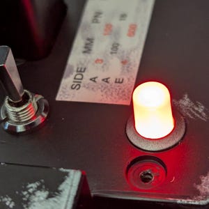 Puede incluir: Primer plano de un panel de control negro con un interruptor de palanca plateado y una luz LED roja. El panel tiene una etiqueta que dice "SIDE A MM PN 3 100 600".