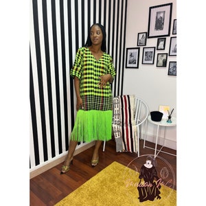 Puede incluir: Un vestido de cuadros verdes y negros con un dobladillo de flecos verdes. El vestido tiene cuello en V y mangas cortas. La modelo lleva el vestido con sandalias doradas.