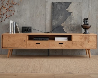 May include: A light-colored wooden TV stand with four doors and two drawers. The stand has a mid-century modern design with angled legs. A small black camera, books, and decorative items are on top. The stand is 72 inches long.