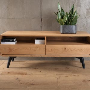 以下が含まれることがあります： 2つの引き出しとオープンシェルフを備えた明るい色の木製テレビ台。丸みを帯びたエッジと黒い脚があります。鉢植えの植物が上に置かれ、本が中にあります。