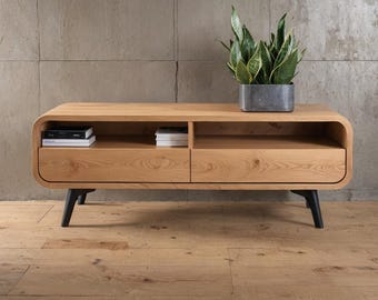 May include: A light-colored wooden TV stand with two drawers and an open shelf. The stand has rounded edges and black legs. A potted plant sits on top, and books are inside the stand.