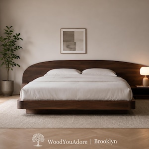 May include: A low-profile dark wood bed frame featuring a wide, arched headboard that extends horizontally. The bed is dressed in plain white bedding with two matching pillows. The text WoodYouAdore | Brooklyn is visible at the bottom.