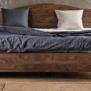 Può includere: Un letto in legno scuro con testiera abbinata. Il letto è in legno scuro e ha un profilo basso. È vestito con un piumino blu scuro e due cuscini, uno blu scuro e uno bianco.