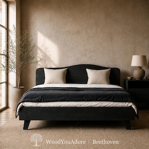 May include: A black wooden bed frame with a curved headboard, featuring a dark wood grain texture. The bed is dressed with a white duvet, a black coverlet, and two beige square pillows. The text WoodYouAdore | Beethoven is at the bottom.