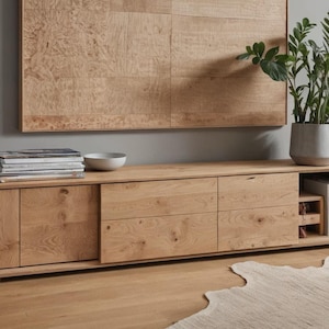 May include: A light oak entertainment unit with drawers and a sliding door. A stack of magazines, a white bowl, and a potted plant are on top. A wooden wall panel is above.