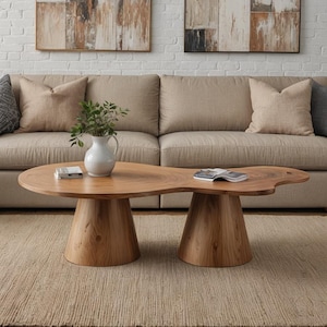 Könnte beinhalten: Einzigartiger Couchtisch aus Holz mit natürlicher, organischer Form. Der Tisch hat zwei konische Sockel, die eine hellbraune, freie Tischplatte tragen. Eine weiße Vase mit Grün und ein Buch liegen auf dem Tisch. Der Tisch befindet sich in einem Wohnzimmer.