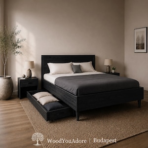 May include: A black wooden bed frame with a tall headboard and a pull-out storage drawer underneath. The bed is dressed with a dark grey duvet, two cream-coloured pillows, and a small grey accent pillow. The frame features a visible wood grain texture.