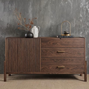 Puede incluir: Aparador de madera marrón oscuro con una puerta de listones verticales a la izquierda y tres cajones a la derecha. El aparador tiene tiradores color latón y está decorado con jarrones y un arco dorado decorativo.