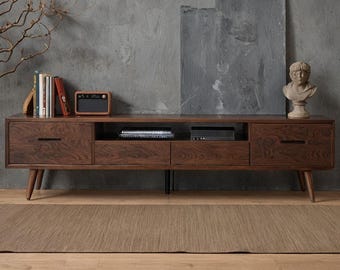 May include: A dark brown wooden media console with two drawers and open shelving. The console has a mid-century modern design with angled legs. A small radio, books, and a bust are on top.