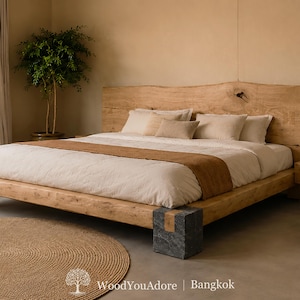 May include: A low-profile wooden bed frame featuring a natural live-edge headboard and a dark gray stone corner support. The bed is dressed in white linens with a tan runner and several beige pillows. Text at the bottom reads WoodYouAdore | Bangkok.