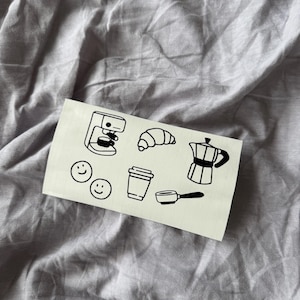 Könnte beinhalten: Ein Aufkleberbogen mit schwarzen Strichzeichnungen von Kaffee-bezogenen Artikeln. Dazu gehören eine Kaffeemaschine, ein Croissant, Smiley-Gesichter, ein To-Go-Becher, eine Espressomaschine und ein Espressokocher. Der Aufkleberbogen liegt auf grauem Stoff.