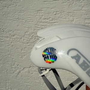 Könnte beinhalten: Weißer Fahrradhelm mit einem holografischen Regenbogen-Aufkleber "CYCLING CLUB". Der Helm hat einen grauen Riemen und der Markenname "ABUS" ist sichtbar. Für Sicherheit konzipiert.