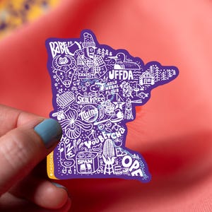May include: A purple sticker in the shape of Minnesota, filled with white illustrations of state icons, including a lumberjack, loon, and the word "SPAM." The sticker is held against a pink background.
