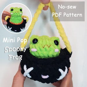 May include: A handmade, green and black crochet frog with a yellow handle. The frog has large black eyes and is sitting in a black pot with white accents. Text on the image reads "Mini Pop Spooky Frog" and "No-sew PDF Pattern".