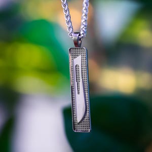 Puede incluir: Collar con colgante plateado, con un colgante rectangular con un diseño de bisturí quirúrgico. El colgante cuelga de una cadena plateada. El fondo es una mezcla borrosa de verde y morado.