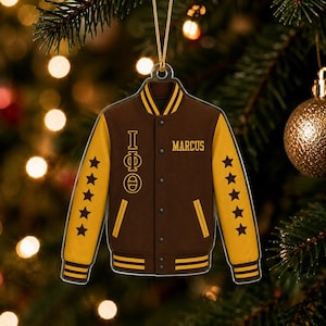 May include: A brown and yellow varsity jacket ornament featuring a gold cord for hanging. The jacket displays the Greek letters Iota Phi Theta vertically, the name MARCUS, and five black stars on each sleeve with striped ribbed trim details.