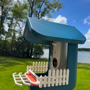 以下が含まれることがあります： 白いピケットフェンスのデザインと内蔵カメラが付いた青いバードフィーダー。フィーダーには赤いトレイと、カメラを見るための透明なプラスチック製の窓があります。