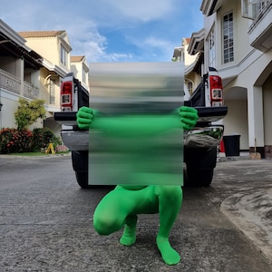 May include: A person in a bright green full-body suit crouches in front of a black pickup truck, holding a rectangular panel with a blurred green and gray design. The background includes houses and a blue sky.