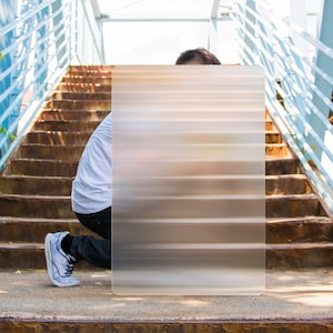 May include: A person kneeling on a concrete surface, partially obscured by a rectangular, blurred image with horizontal lines. The person wears a white t-shirt, black pants, and gray sneakers. The background features a staircase and a blue railing.