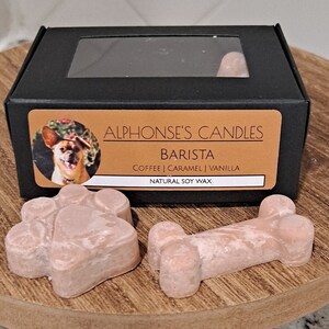 May include: A black box with a clear window showing two wax melts in the shape of a paw print and a bone. The box has a label that says "Alphonse's Candles" and "Barista" with the scent description "Coffee | Caramel | Vanilla" and "Natural Soy Wax".