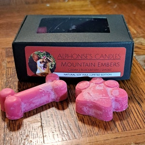 Op de afbeelding: Een zwarte doos met een transparant venster dat twee roze wax melts laat zien in de vorm van een bot en een poot. De doos heeft een label met de tekst "Alphonse's Candles Mountain Embers Cedar | Blackberry | Smoke Natural Soy Wax | Limited Edition".