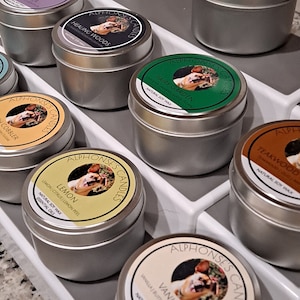 May include: A collection of small, silver metal tins of Alphonse's Candles, each with a different scent. The labels feature a dog illustration and scent names like "Lemon," "Vanilla," and "Sacred Amber." The candles are arranged on a white tray.