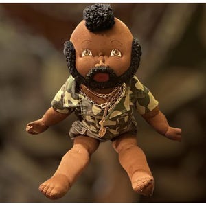 May include: A fabric doll with dark brown skin, a black mohawk hairstyle, and a matching black beard. The doll wears a green and brown camouflage-patterned short-sleeved shirt and shorts, accessorized with multiple gold-toned metal chain necklaces.