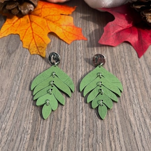 May include: A pair of green leaf-shaped earrings with silver accents. The earrings are made of wood and have a layered design.