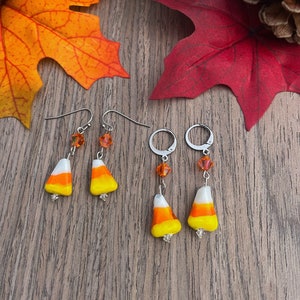 Puede incluir: Un par de pendientes colgantes con un diseño de caramelo de maíz con detalles en blanco, naranja y amarillo. Los pendientes tienen un aro plateado y un dije de cristal.
