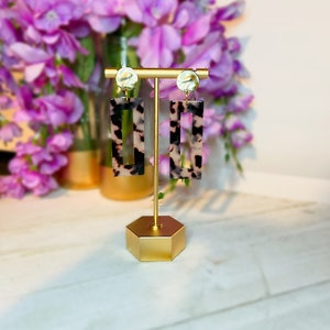 May include: A pair of tortoise shell rectangle earrings with gold accents. The earrings are on a gold stand.