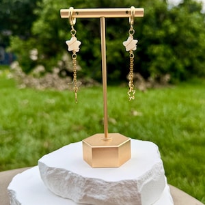 Könnte beinhalten: Ein Paar goldfarbene Hängeohrringe mit einem kleinen weißen Sternanhänger, der mit Golddraht umwickelt ist und an einer zarten Goldkette hängt, die in einem kleinen, durchbrochenen Goldstern endet. Die Ohrringe hängen an einem T-förmigen Schmuckständer.