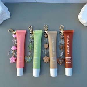 May include: Four tubes of Lakerain Summer Special lip balm butter in coral, light green, beige, and brown. Each tube features a gold-tone lobster clasp keychain with decorative charms including hearts, stars, bows, flowers, and butterflies.