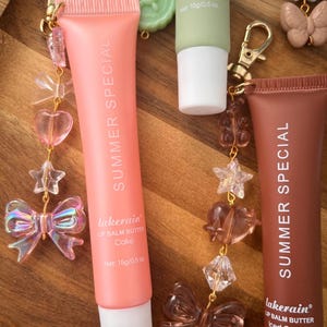 以下が含まれることがあります： ピンク、緑、茶色のリップバームチューブ。テキスト「SUMMER SPECIAL」と「lakerain LIP BALM BUTTER」入り。各チューブには、リボン、ハート、星の装飾チャームが付いています。リップバームはそれぞれ15gです。