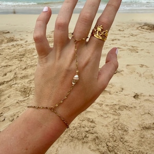 May include: A gold-colored hand chain and ring set. The ring features a palm tree design. The hand chain connects to a small seashell. The background shows a sandy beach and ocean.
