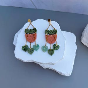 Könnte beinhalten: Ein Paar Hängeohrringe mit goldenem Stecker und Kettendesign. Jeder Ohrring zeigt einen terrakottafarbenen, halbkreisförmigen Pflanztopf mit Gittermuster, der mit kleinen grünen Blättern und drei hängenden blattförmigen Anhängern verziert ist.