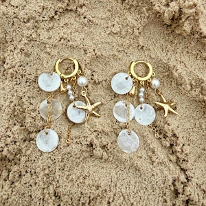 May include: A pair of gold hoop earrings with dangling white shell discs, small pearls, and gold starfish charms. The earrings are displayed on a sandy surface, evoking a beach theme.