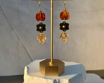 Pode incluir: Um par de brincos pendentes com um pingente de abóbora laranja, um componente preto em forma de flor com centro dourado e um pingente de folha dourada texturizada. Os brincos estão suspensos em ganchos dourados num suporte de metal.