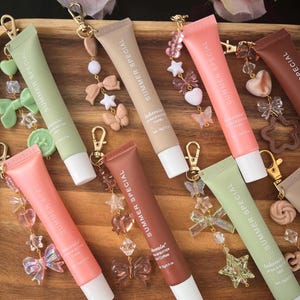 May include: A selection of lip balm tubes in shades of pink, beige, and green, labelled "SUMMER SPECIAL". Each tube is embellished with a charm featuring bows, stars, and butterflies. The lip balms are presented on a wooden tray.