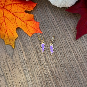 May include: A pair of gold hoop earrings with purple lightning bolt charms. The earrings are on a wooden surface with fall leaves.