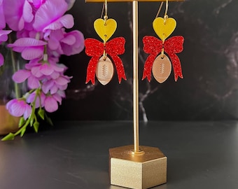 May include: A pair of earrings with a football theme. Each earring features a yellow heart, a red glitter bow, and a football-shaped charm. The earrings are displayed on a gold stand against a dark background.